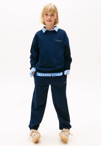 Navyblauwe sweatpants en een bijpassende crew neck sweatshirt met logo, gelaagd over een lichtblauw gestreept shirt. Materiaal is zacht, casual pasvorm.