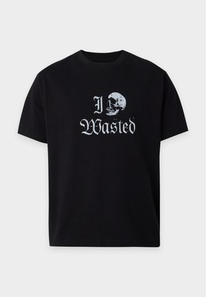 Млад мъж, носещ черна тениска с текст "I Wasted" и графика на череп, съчетана със светлосиви спортни панталони с малко лого отляво.