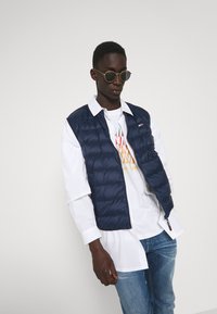 Chaleco de plumas azul marino con cremallera, que presenta un diseño acolchado. Llevado sobre una camisa blanca con un estampado gráfico y combinado con unos jeans de color azul claro.