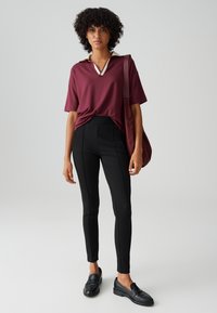 Maglietta polo bordeaux con colletto bianco, abbinata a pantaloni neri aderenti con cuciture laterali. Il modello indossa mocassini neri e porta una borsa bordeaux.