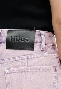 Πίσω όψη τζιν από ντένιμ σε χρώμα λεβάντας με μαύρη δερμάτινη ετικέτα με ανάγλυφο το λογότυπο "HUGO", φορεμένα με μαύρο τοπ.