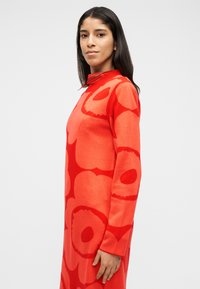 Γυναίκα με ζωηρό κόκκινο φόρεμα με μεγάλα αφηρημένα κυκλικά σχέδια, σε πλάγια στάση μπροστά σε απλό φωτεινό φόντο, κοιτάζοντας την κάμερα.