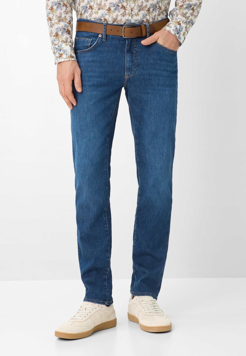 Uomo che indossa jeans aderenti blu, cintura di pelle marrone, camicia a maniche lunghe a fiori e sneakers bianche, in piedi con una mano in tasca.
