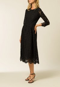 Svart spets midi-klänning med långa ärmar, figurnära överdel, utsvängd kjol och scalloped nederkant. Bärs med svarta sandaletter med remmar.