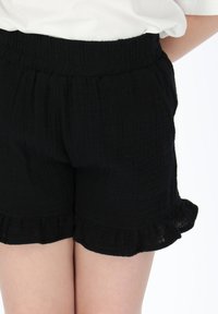 Schwarze, gekräuselte Shorts mit elastischem Bund und gerüschtem Saum. Der Stoff wirkt leicht und strukturiert.