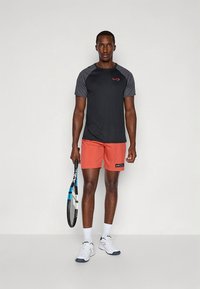 Kortärmad svart och grå sporttröja med en röd logotyp, kombinerad med korallfärgade shorts. Håller i en tennisträning, bär vita skor.