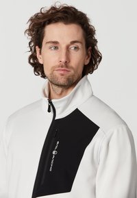 Vit zip-up jacka med en svart panel på bröstet, har hög krage och logodetalj. Materialet verkar vara texturerat och hållbart.