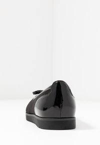 Zapato de ante negro con un tacón de charol brillante. Cuenta con una punta redonda y un detalle de cinta negra en el lateral. Suela plana de goma.