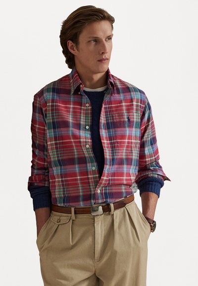 Rutigt skjorta i röd, grön och blå bomullstyg, med knäppning på kragen, bröstficka och rullade ärmar. Stylad med khaki-byxor.