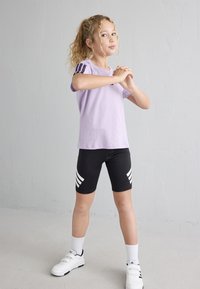 Vaalean violetti lyhythihainen paita, jossa on tumman violetit raidat olkapäissä, yhdistettynä mustiin pyöräilyshortseihin, joissa on valkoisia raidallisia yksityiskohtia. Valkoiset kengät.