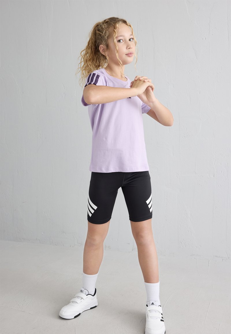 Vaalean violetti lyhythihainen paita, jossa on tumman violetit raidat olkapäissä, yhdistettynä mustiin pyöräilyshortseihin, joissa on valkoisia raidallisia yksityiskohtia. Valkoiset kengät.