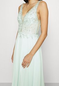 Mátově zelené večerní šaty s beaded korzetem, výstřihem do V a průhlednými vyšívanými detaily na vrchu. Plynulá sukně s vrstvenými látkami.