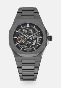 Montre squelette grise avec boîtier octagonal, bracelet en acier inoxydable et mouvement mécanique exposé. Marqueurs d'heure et aiguilles blancs.
