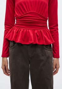 Rød genser med lange ermer, innsvingt midje og vid peplum-detalj, kombinert med mørkebrune bukser med vide ben. Bomullsblanding med myk tekstur.