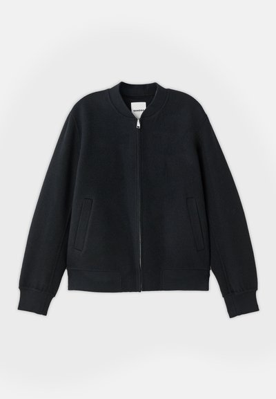 Must must jacket, mis tekstureeritud kangast, millel on tõmblukk ees, soonilised mansetid ja kaks külgtaskut. Lihtne, minimalistlik disain.
