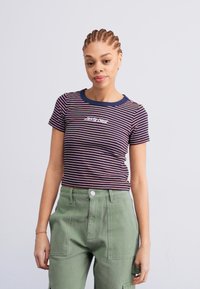 T-shirt a righe blu navy, rosso e bianco. Materiale in cotone, maniche corte, scollatura a girocollo ribattuta con testo ricamato. Indossato con pantaloni verde oliva.