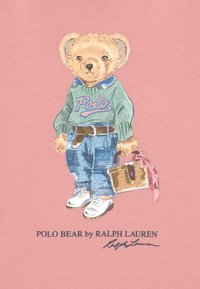 Illustration av en björn som bär en grön "Polo"-tröja, blå jeans och vita sneakers, hållande en vävd väska med en rosa scarf. Bakgrunden är rosa.