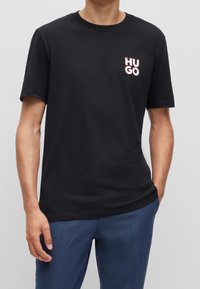Man in een zwart T-shirt met korte mouwen en het "HUGO"-logo op de linkerborst, in blauwe broek met één hand in zijn zak.