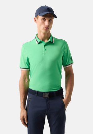 Mężczyzna w jaskrawozielonej koszulce polo z paskowanym kołnierzykiem i rękawami, granatowych spodniach, pasku i granatowej czapce na białym tle.