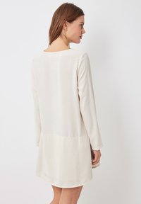 Vestido de manga larga en color crema, confeccionado con una tela suave y ligera. Presenta un escote alto y un dobladillo ligeramente acampanado, con un detalle de costura en la parte posterior.