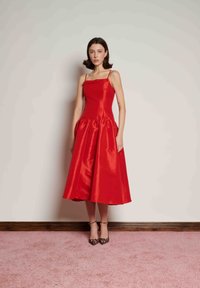 Vestido de cetim vermelho com corpete justo e saia evasê abaixo do joelho. Alças finas e ligeiro brilho. Usado com sapatos de salto estampados em padrão de leopardo.