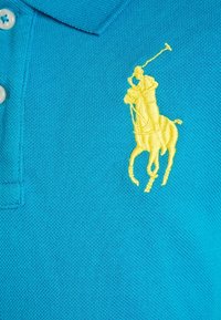 Pikétröja i klart turkos bomull med en texturerad yta, som har en framträdande gul broderad logotyp av en häst och ryttare på bröstet.