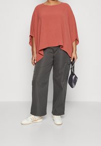 Locker sitzendes, rostfarbenes Oberteil mit weitem Ärmel und einem abfallenden Saum, kombiniert mit dunkelgrauen Cargo-Hosen und weißen Sneakern. Hält eine kleine schwarze Tasche in der Hand.