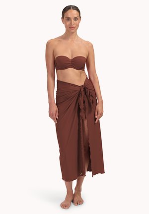 Kobieta stojąca boso, ubrana w brązowy, bezramienny top bandeau oraz dopasowaną sarongę zawiązaną w talii, z przednim rozcięciem.