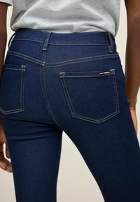 Jeans in denim blu scuro con un design aderente, caratterizzati da cuciture a contrasto e tasche posteriori. La vita è liscia e di media altezza.