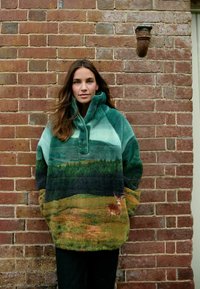 Pull en polaire vert et bleu avec un imprimé de paysage présentant des arbres et un cerf, col montant et poches avant, posé contre un mur en briques.