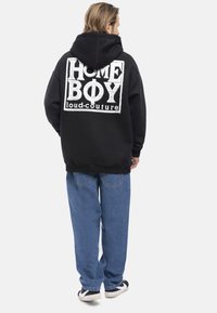 Felpa nera con un grande grafiche bianco che recita "HOME BOY loud-couture" sulla schiena. Abbinata a pantaloni in denim blu e sneakers nere.