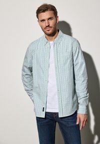 Zeleno-bílá kostkovaná košile s límcem a dlouhými rukávy, přes bílou tričko, doplněná tmavě modrými džíny.