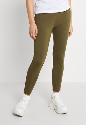 Person wearing olive green slim-fit pants, white socks, chunky white sneakers, and a white top, standing on a plain light grey floor.