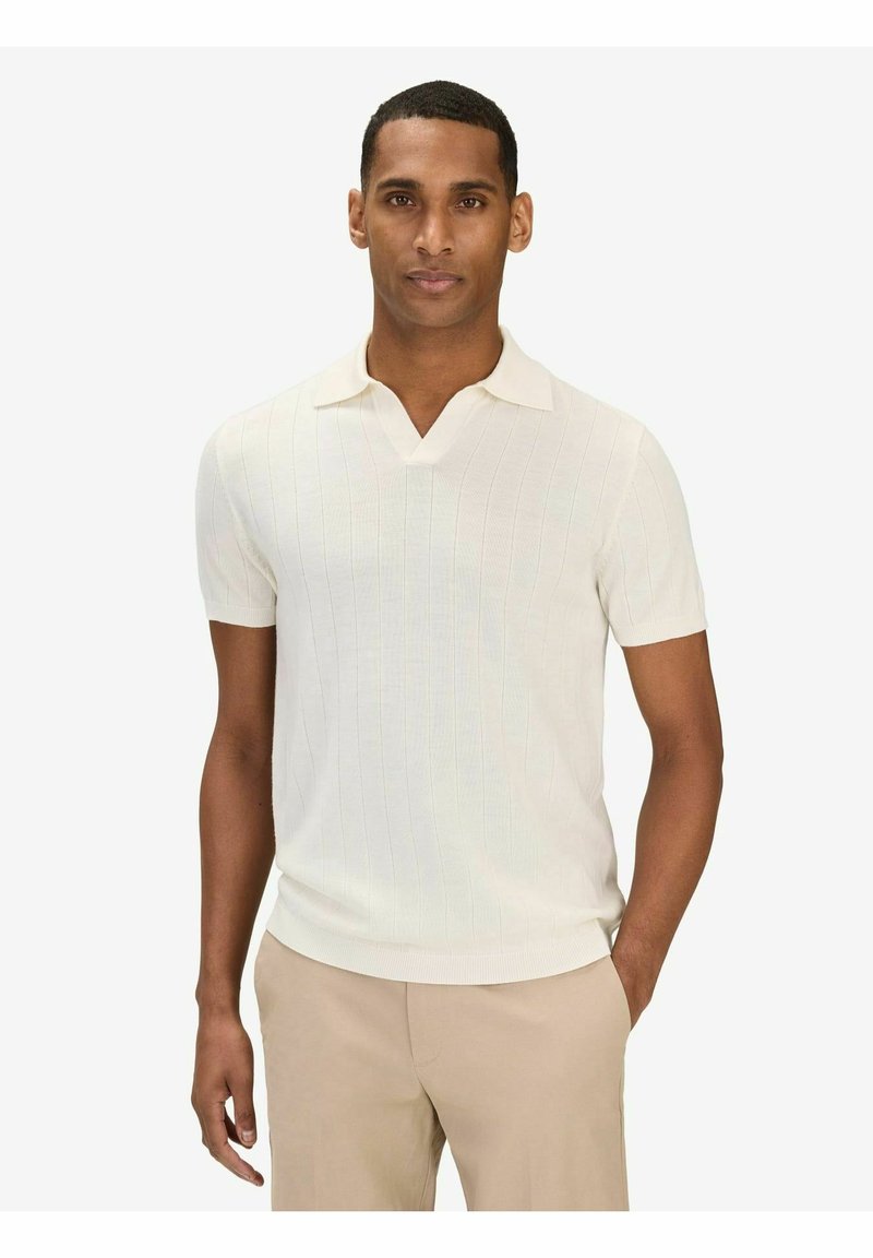 Homme portant un polo en maille blanc à manches courtes et un pantalon beige, debout avec une main dans la poche devant un fond uni.