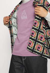 Camicia aperta davanti con un motivo grafico multicolore, indossata sopra una maglietta semplice lavanda con logo bianco stampato. Pantaloni grigio scuro visibili.