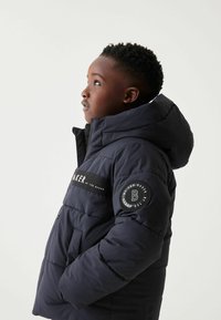 Doudoune bleu marine avec capuche, ornée d'écussons de logo, de coutures horizontales et d'une fermeture éclair à l'avant. Le matériau semble lisse et isolé.