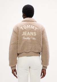 Béžová, nadýchaná kožušinová bunda s límcom. Na chrbte je biele vyšívanie s nápismi "TOMMY JEANS" a "Roadtrip USA". Hladké manžety.