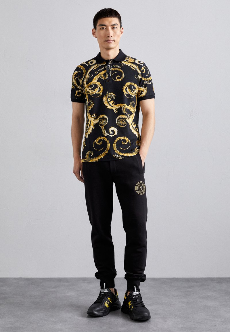 La camiseta polo presenta una base negra con patrones dorados en espiral, combinada con pantalones de chándal negros que exhiben un logo dorado. Zapatillas negras completan el conjunto.