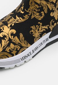 Svart sko med ett guldigt barockmönster, elastisk ovandel, vit sula med varumärket "VERSACE JEANS COUTURE" och en texturerad yta.