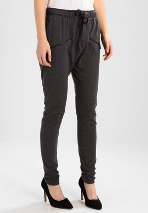 Osoba ubrana w ciemnoszare spodnie dresowe z zamykanymi na zamek kieszeniami z przodu oraz czarne buty na wysokim obcasie, stojąca na jednolitym tle.