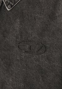 Svart denimjacka med en uttalad texturerad yta och en broderad logotyp på tyget. Kragn_designen ger struktur åt plagget.