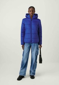 Zila polsterēta jaka ar kapuci, ar priekšējo rāvējslēdzēju un quilted dizainu. Pāris ar platām zilām kravas biksēm un melnu apavu.