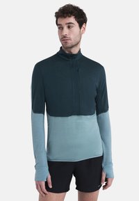 Camiseta deportiva de manga larga con cuello con cremallera, diseño a bloques de color en verde azulado oscuro y azul claro, textura suave y silueta ajustada.