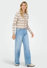 Vrouw die loopt in een beige en witte gestreepte button-up trui, lichtblauwe wijde jeans en crèmekleurige loafers op een witte achtergrond.