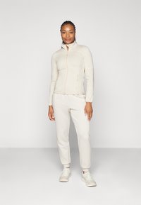 Hellbeige Reißverschlussjacke mit strukturierten Einsätzen, kombiniert mit passenden schmal zulaufenden Hosen. Das Model trägt helle Sneakers und präsentiert einen lässigen, sportlichen Stil.
