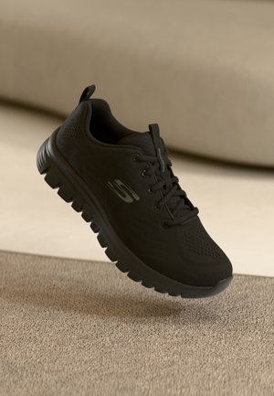 Czarny but sportowy Skechers z grubą podeszwą z bieżnikiem, z przodu na sznurowadła i logo po boku, przedstawiony na beżowym tle.