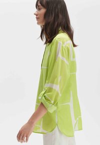 Lime green blouse with white geometric patterns, long sleeves rolled up, light fabric, button detail on sleeves, slightly curved hemline.