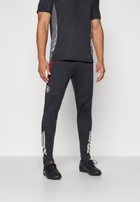 Sorte atletiske bukser med en tætsiddende design, der har hvide striber på siderne og et logo på låret. Fremstillet af strækbart stof.