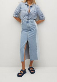 Chemise en denim bleu clair avec poches à boutons, associée à une jupe taille haute longueur genou avec une fente à l'avant et un panneau latéral contrastant. Sandales bleues.