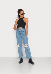 Top cropped preto com alças, jeans de cintura alta azul-claro com grandes rasgos desgastados e botas pretas de salto grosso. Óculos de sol como acessório.